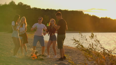 Company-of-young-people-are-dancing-in-shorts-and-t-shirts-around-bonfire-on-the-open-air-party-with-beer.-They-are-enjoying-the-summer-evening-at-sunset-near-the-forest.